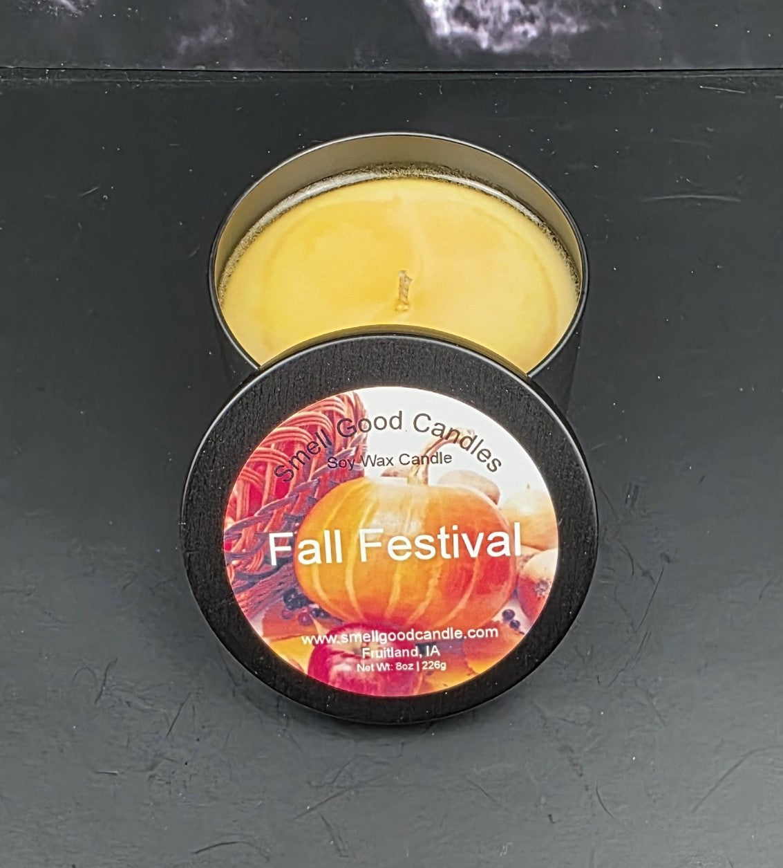 Scented candle in a tin labeled 'Fall Festival' on a dark surface
